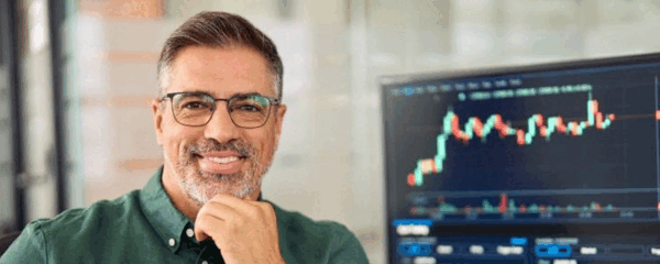 A man wearing glasses and a green shirt smiles while standing before a computer screen, indicative of a forex broker.