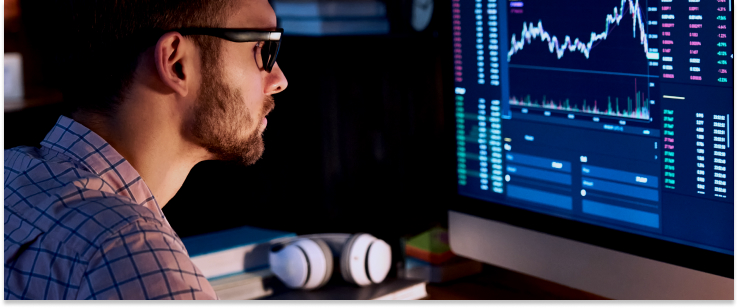 A man with glasses analyzes stock market graphs on a computer screen, focused on forex trading.
